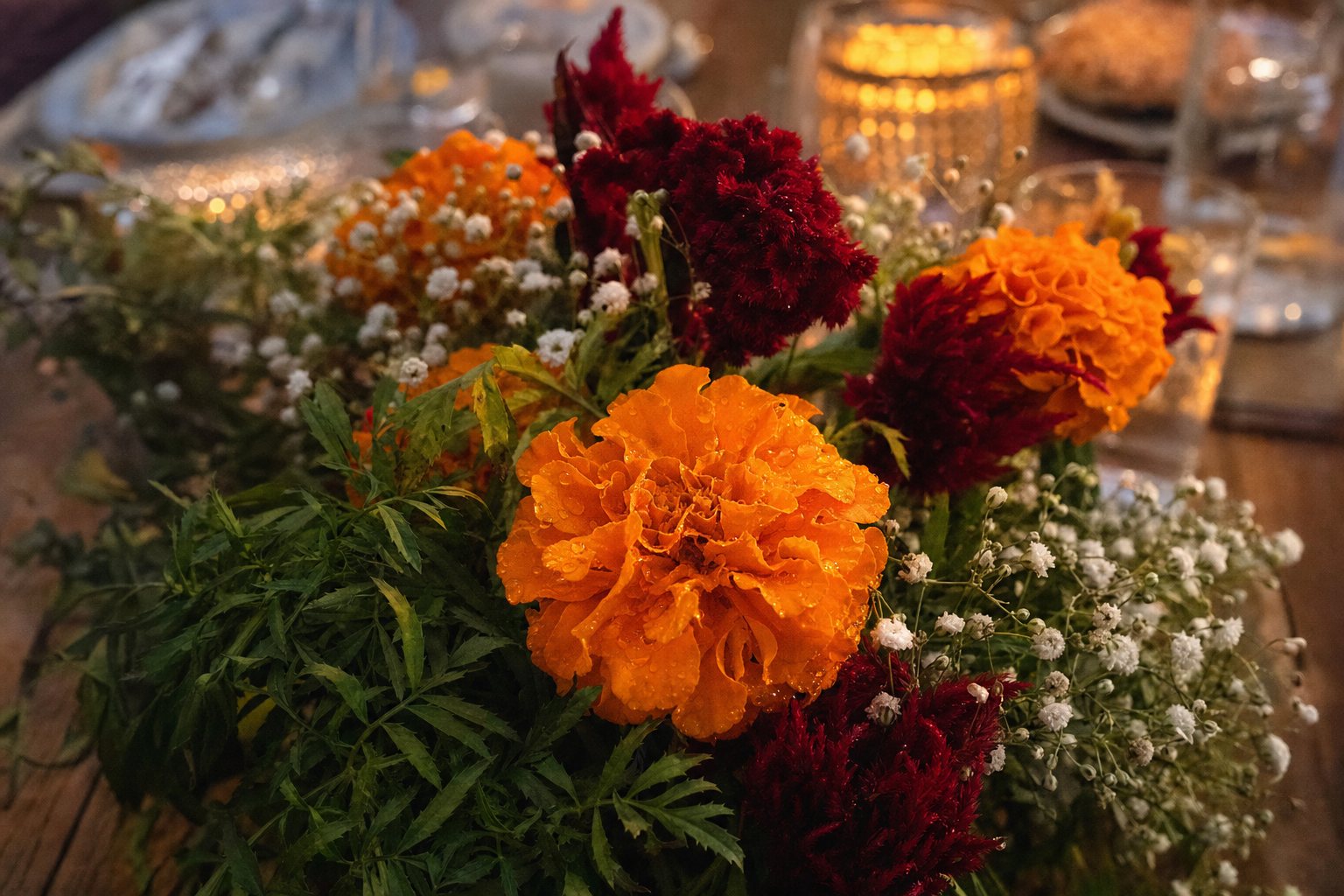 Bright orange and gold marigolds with red celosia and white baby's breath spread across a sunlit wooden table