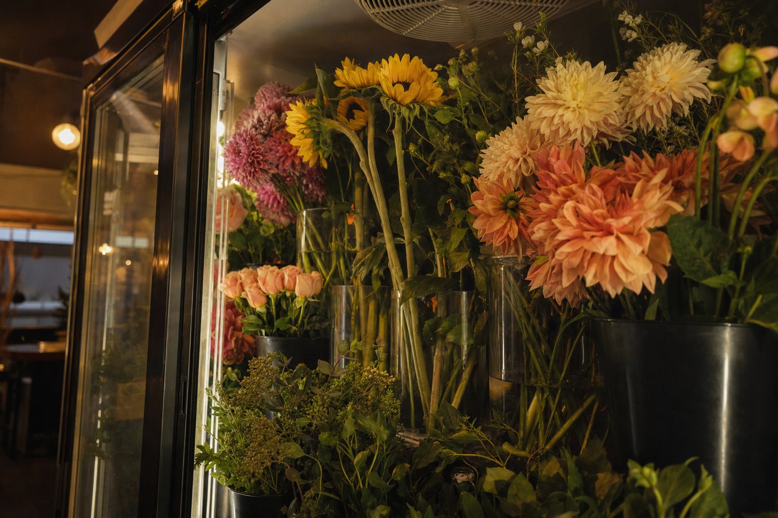 Glass-door florist cooler stocked with buckets of dahlias, sunflowers, roses and greenery in clear water