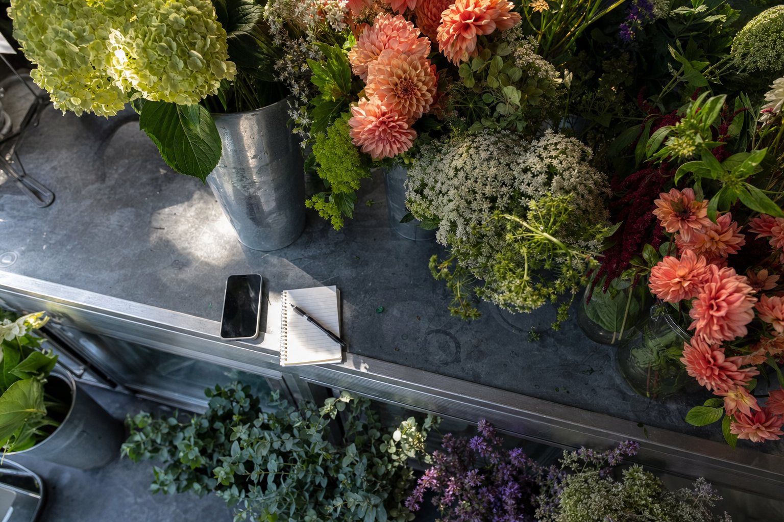 Sunlit flower shop counter with a phone, notepad, and buckets of bright seasonal flowers